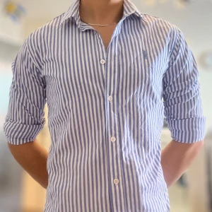 Blåvit randig skjorta från Ralph Lauren - Snygg skjorta från Ralph Lauren i blått och vitt med vertikala ränder. Klassisk krage, knappar framtill och långärmad design. Tillverkad i mjuk bomull som känns skön mot huden. Perfekt för dig som gillar stilrena och fräscha plagg med ikonisk logga på bröstet.