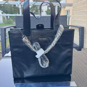 Ny Svart handväska / crossover  - Ny stilren svart handväska med raka linjer och rejäla handtag. Väskan har en avtagbar kedjerem i guld och ett lila innerfoder med dragkedja på innerfacket. Har magnetlås. Får plats en skoldator. Finns på Hägerstensåsen i Stockholm. 