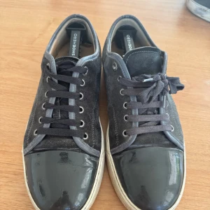 Svarta sneakers från Lanvin med lacktå - Säljer ett par svarta Lanvin sneakers med snygg lackad tå och ovandel i mocka. Skorna har rund tå, platt sula och klassisk snörning. Grå detaljer längs kanterna och vita sulor ger en stilren vibe. Perfekta för dig som gillar exklusiva och coola sneakers. Dock så har dom inte samma snören det ena är lite mörkare än den andra dom har också ett litet hål på botten.