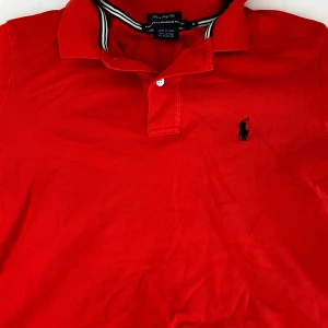Röd pikétröja Ralph Lauren Golf S - Riktigt snygg röd pikétröja från Ralph Lauren Golf i klassisk modell med svart broderad logga på bröstet och AIGA-märke på ärmen. Tröjan har krage med vit-svarta ränder och är gjord i mjuk bomull med lite stretch. Perfekt för golf eller chill dagar.