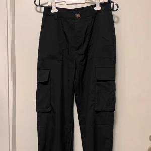 Svarta cargopants med fickor - Svarta cargopants med stora sidofickor och knappstängning i midjan. Byxorna har en loose passform och är perfekta för en chill och streetig stil. Materialet känns lätt och bekvämt, och de har klassiska cargodetaljer som ger en cool vibe.