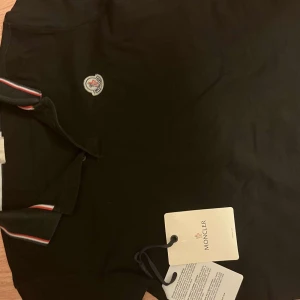 Svart Moncler polo med logga - Svart t-shirt från Moncler med klassisk logga på bröstet och snygga röd-vita detaljer på kragen. T-shirten har korta ärmar och är gjord i mjuk bomull som känns skön mot huden. Perfekt för dig som gillar stilrena märkesplagg med en sportig touch. Sitter som S-M