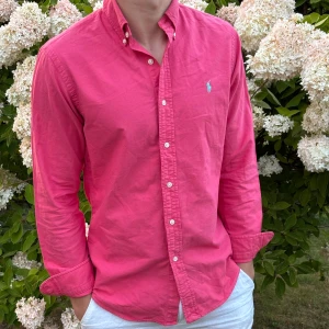 Rosa skjorta från Polo Ralph Lauren - Snygg rosa skjorta från Polo Ralph Lauren i slim fit. Skjortan har klassisk krage, knappar hela vägen och lång ärm. Tillverkad i mjuk bomull och har den ikoniska broderade loggan på bröstet. Perfekt för dig som gillar stilrena och färgstarka plagg. Modellen är 182cm och 73kg 