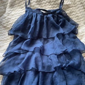 Blå volanglinne från Lindex Kids - Supersöt blått linne från Lindex Kids med flera lager volanger och glansiga paljetter runt bandet. Toppen har en luftig och flowy känsla. Materialet är lätt och ger snygg rörelse när du går.