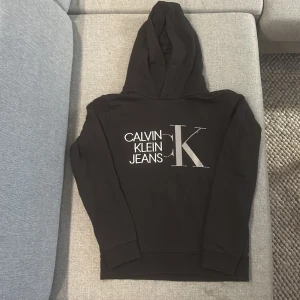 Svart Calvin Klein Jeans hoodie - Svart hoodie från Calvin Klein Jeans med stor CK-logga och vit text på bröstet. Tröjan har huva och långa ärmar, tillverkad i mjuk bomull. Perfekt för dig som gillar stilrena och klassiska streetwear-plagg med tydlig branding. 150-160cm. Inga fel i bra skick.