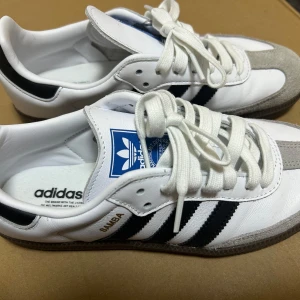Adidas Samba sneakers vit/svart 38 2/3 - Adidas Samba sneakers i klassisk stil med vit ovandel i skinn, svarta ränder på sidorna och grå mockadetaljer vid tån. Skorna har snörning, platt sula och rund tå. Blå Adidas-logga på plösen och gummisula för extra grepp. Perfekt för dig som gillar retrovibe och streetstyle. Skorna har använts i 2 gånger bara för att de var för små. De är i väldigt bra skick. 