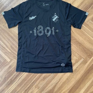 Svart AIK fotbollströja Nike M - AIK fotbollströja i svart med trycket '1891' på bröstet och klubbmärket. Tillverkad i Dri-FIT material som andas, perfekt för träning eller match. Kortärmad modell med diskreta detaljer på ärmarna och Nike-logga. Snygg och stilren design för AIK-supportern.