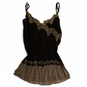 Mörk/ljusbrun och guldig linnetopp som är räfflad med blommig spets. - Supersöt brun linnetopp med tunna axelband och vacker spetsbrodyr i guld längs urringningen och nedtill. Toppen har plisserad, luftig design och volangkant nertill som ger en romantisk vibe. Perfekt att styla med jeans eller kjol för en cool look.