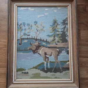 Tavla med motiv av en älg i naturen, broderad  - Upptäck en unik broderad tavla med motiv av en älg vid en sjö, omgiven av träd och natur. Perfekt för dig som gillar djur och natur. Fint skick. Bredd 26 cm, längd 34 cm. 