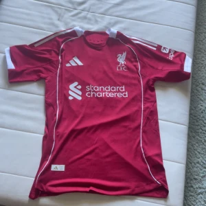 Liverpool hemmatröja 25/26 med Virgil #4 på ryggen - Liverpool FC 25/26 matchtröja med Virgil och nummer 4 på ryggen. Tröjan är röd med vita detaljer, korta ärmar och klassiska Adidas-ränder på axlarna. Materialet är lätt och ventilerande polyester, perfekt för fotboll. LFC-logga och sponsortryck på bröstet. Aldrig använd bara testad en gång. Storlek M och pris kan diskuteras.