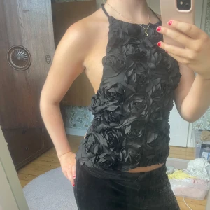 Svart blomstertopp med öppen rygg - Supersnygg svart halterneck topp med 3D-blommor över hela framsidan. Toppen har öppen rygg och knyts med tunna band både i nacken och ryggen. 