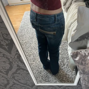 Blå jeans från Fornarina med dragkedja - Säljer ett par blå jeans från Fornarina med coola detaljer som snedställd bakficka med dragkedja. Jeansen har normal passform och är tillverkade i bomull med lite stretch för extra komfort. Perfekt för dig som gillar unik design och streetstyle.