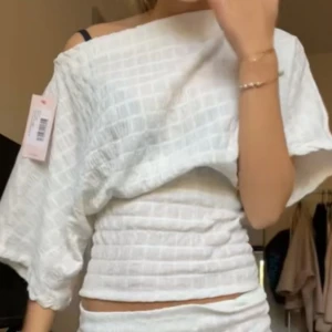 Vit offshoulder blus med struktur - Säljer en vit blus med offshoulder och trekvartsärm. Toppen har ett strukturerat rutmönster och är croppad med smockad midja som ger en snygg passform. Perfekt för dig som gillar trendiga detaljer och ljusa färger.