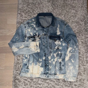 Blå jeansjacka med blekta detaljer - Säljer en snygg blå jeansjacka med coola blekta splash-effekter över hela jackan. Klassisk modell med krage, knappar framtill och fickor på bröstet. Perfekt för dig som gillar streetstyle och vill sticka ut med en unik look.