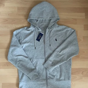 Grå hoodie från Polo Ralph Lauren - Snygg grå hoodie från Polo Ralph Lauren med dragkedja och huva. Tröjan har långa ärmar, snörning vid huvan och klassisk Polo-logga broderad på bröstet. Perfekt för chill dagar och enkel att matcha med jeans eller joggers.