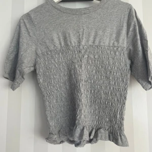 Grå ribbad topp med volangkant - Supersöt grå topp med ribbad struktur på nedre delen och volangkant längst ner. Kortärmad och rund halsringning, perfekt för dig som gillar en chill och trendig stil. Materialet känns mjukt och skönt mot huden.