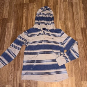 Randig hoodie från Polo Ralph Lauren - Snygg hoodie från Polo Ralph Lauren i vitt och blått med randigt mönster. Tröjan har huva med snörning, knappar vid halsen och en broderad logga på bröstet. Långärmad och tillverkad i mjuk bomull, perfekt för chill dagar.