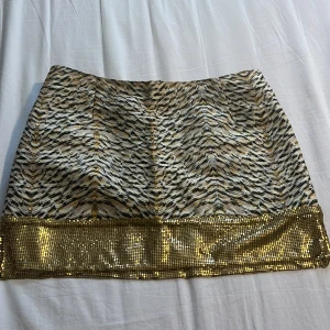 Kort kjol med guldpaljetter - Säljer en kort kjol med ett coolt zebramönster i svart, beige och guld. Nederkanten är täckt av glittriga guldpaljetter som ger extra bling. Kjolen har en rak passform och är perfekt för dig som gillar att sticka ut med din stil.