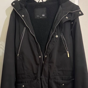 Svart parkajacka med teddyfoder  - Svart parkajacka från 157 i storlek M, med mjukt teddyfoder och stor huva. Jackan har flera dragkedjor och tryckknappar framtill, samt praktiska fickor. Perfekt för kalla dagar med sin varma och fluffiga insida. Snygg och enkel design som funkar till många stilar.