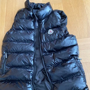 Svart dunväst från Moncler - Snygg svart dunväst från Moncler med glansig finish och klassisk logga på bröstet. Västen har hög krage, två fickor med dragkedja och quiltad design. Perfekt att slänga över en hoodie när det är kyligt ute. Innerfoder med coolt serietryck.