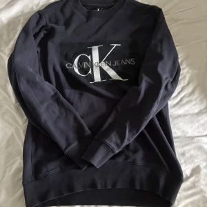 Mörkblå sweatshirt från Calvin Klein - Snygg mörkblå sweatshirt från Calvin Klein Jeans med stort CK-logotryck på bröstet. Tröjan har rund hals, långa ärmar och ribbade muddar vid ärmslut och nederkant. Perfekt för dig som gillar stilrena och klassiska märkesplagg.