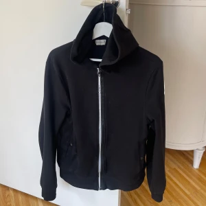 Svart Moncler hoodie med dragkedja - Svart hoodie  från Moncler med hög krage och tydlig logga på ärmen. Hoodien har hel dragkedja framtill, två fickor med knappstängning och är tillverkad i mjukt bomullsmaterial. Snygg och enkel design som passar till många olika stilar.