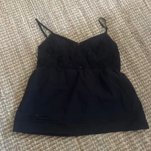 Svart broderad topp med knappar - Svart topp med tunna axelband och broderade blommor på överdelen. Toppen har små knappar framtill och en volang nedtill. 