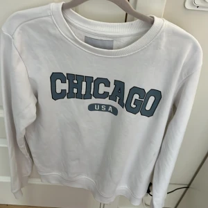 Vit sweatshirt med Chicago-tryck 157 - Vit sweatshirt från 157 med stort blått CHICAGO USA-tryck på bröstet. Tröjan har rund hals, ribbade muddar och är tillverkad i mjuk bomullsmix med fleece på insidan. Perfekt för dig som gillar college-vibe och streetstyle.