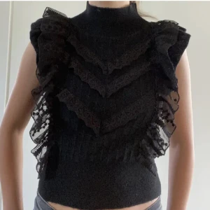 Svart volang topp med spetsdetaljer - Supersnygg svart topp med höghalsad design och volanger i lager på framsidan. Spetsdetaljer längs volangerna ger en cool och unik look. Toppen är ärmlös och croppad, perfekt för att sticka ut med en edgy vibe.