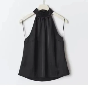 Svart ärmlös halterneck blus - Supersnygg svart blus med halterneck och hög krage. Den är ärmlös och har en lätt glansig finish som ger en lyxig känsla. Perfekt att styla med jeans eller kjol för en cool look. Materialet känns mjukt och följsamt mot huden.