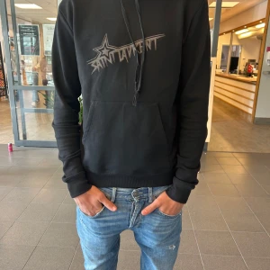 Svart Saint Laurent hoodie med tryck - Svart hoodie från Saint Laurent med stort grafiskt tryck på bröstet. Klassisk huva med snörning och magficka framtill. Långärmad och i mjukt bomullsmaterial, perfekt för en chill och stilren look.