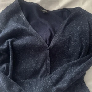 Glittrig marinblå kofta från Alice Bizous - Snygg marinblå kofta från Alice Bizous med glittriga detaljer. V-ringad och knäpps med tre knappar framtill. Mjuk och stretchig kvalitet, perfekt för att addera lite extra stil till din outfit. Långärmad och med ribbad kant nedtill.