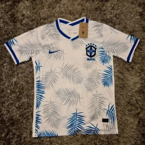 Vit Brasil fotbollströja  - Snygg Brasil fotbollströja från Nike i vit med blå detaljer och palmbladsmönster över hela tröjan. Kortärmad med blått v-ringad krage och blå kant vid ärmslut. Materialet är lätt och andas, perfekt för match eller träning. Brasil-logga och Nike Swoosh på bröstet.
