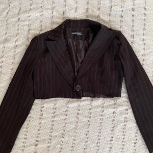 Brun croppad kavaj från Forget Me Not - Snygg brun croppad kavaj med smala vita ränder från Forget Me Not. Klassisk krage, lång ärm och en knapp framtill. Perfekt för dig som vill ha en trendig och stilren look med en twist. Tunn fodring och figurnära passform.