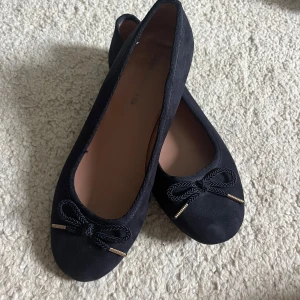 Mörkblåa ballerinaskor - Stilrena marinblåa ballerinaskor från Tamaris i mocka-look med rund tå och dekorativ rosett framtill med gulddetaljer. Lite repor men annars bra skick💕Är borta just nu så kan skicka på måndag!