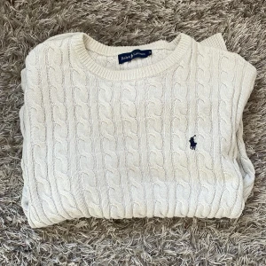 Vit kabelstickad tröja Ralph Lauren - Kabelstickad tröja från Ralph Lauren i klassisk vit färg. Tröjan har rund halsringning, lång ärm och den ikoniska broderade loggan på bröstet. Perfekt för lager-på-lager och en clean look. Tillverkad i mjuk bomull för skön känsla.