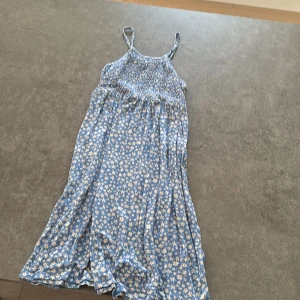 Blommig blå sommarklänning från Kiabi - Supersöt blå klänning från Kiabi med vita och gula blommor. Smockad upptill och tunna axelband ger en luftig känsla. Perfekt för varma dagar med sitt lätta bomullsmaterial och flowiga passform.