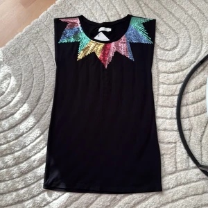 Svart paljettopp från att. - Svart ärmlös topp från att. med färgglada paljetter i ett stjärnformat mönster över axlar och bröst. Paljetterna skiftar i grönt, blått, gult, rött och rosa och ger en riktigt cool statement-look. Toppen är i mjukt material och har rund halsringning.