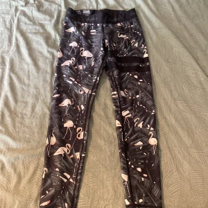 Svarta leggings med flamingomönster STRONGER - Snygga svarta leggings från STRONGER med tropiskt mönster av rosa flamingos och gröna blad. Hög midja och stretchigt material som sitter tajt och bekvämt. Perfekta för träning eller chill dagar. Elastisk midja med logga.