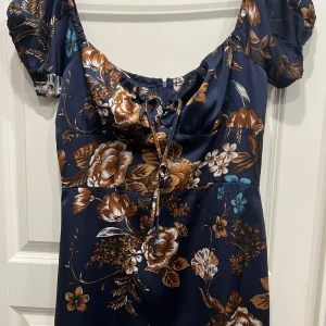 Blommig marinblå klänning från Boohoo - Supersöt marinblå kortklänning från Boohoo med bruna och vita blommor, puffärmar och snörning framtill. Perfekt för dig som gillar romantiska vibes och vill sticka ut lite extra. Lätt och skön att bära, passar perfekt till varmare dagar.