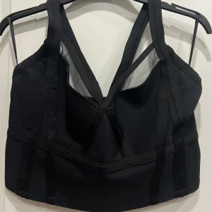 Svart korsettopp från Bubbleroom - Snygg svart korsettopp från Bubbleroom med breda axelband och markerade sömmar. Toppen har en cool v-ringning och meshdetaljer framtill som ger en edgy vibe. Perfekt att styla med höga jeans eller kjol för en trendig look.