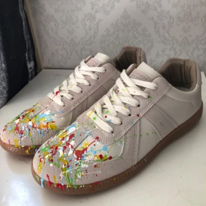 Vita Maison Margiela Gats - Helt nya Maison Margiela Gats med beige och vita paneler i skinn och mocka, samt coola färgstänk i rött, blått, gult och grönt. Dock så är de inte äkta (vilket inte är synligt förutom en liten text under logot som täcks med skosnörerna). Perfekt för dig som vill sticka ut med en konstnärlig vibe.