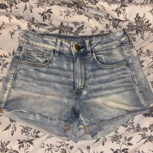 Ljusblå jeansshorts från American Eagle - Snygga ljusblå jeansshorts från American Eagle med fransig kant och klassisk femficksdesign. Shortsen har slitna detaljer och en bekväm midwaist-passform. Perfekta för varma dagar och enkel att matcha med allt.