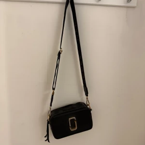 Svart axelväska från Marc Jacobs - Snygg svart axelväska från Marc Jacobs med justerbart axelband och guldiga metalldetaljer. Väskan har en rektangulär form, dubbla dragkedjor och en ikonisk logga framtill. Perfekt storlek för mobil, plånbok och andra essentials.