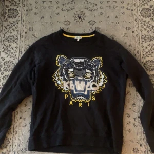 Svart Kenzo tröja med tigertryck - Svart långärmad tröja från Kenzo med ikoniskt broderat tigerhuvud i blått, vitt och gult framtill. Rund halsringning och mjuk bomullskänsla. Perfekt statement-plagg för dig som gillar streetstyle och vill sticka ut.