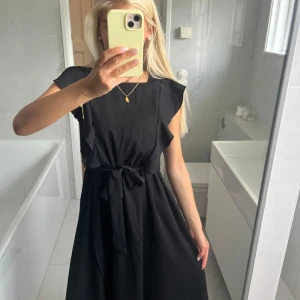 Svart kortklänning med knyt i midjan - Snygg svart klänning med korta ärmar och volangdetaljer längs sidorna. Knytband i midjan som ger en fin siluett. Perfekt för dig som gillar en enkel men stilren look. Klänningen har rund halsringning och är lätt utsvängd nertill.