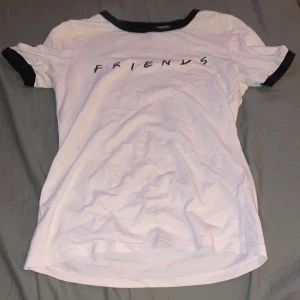 Vit Friends t-shirt från H&M Divided - Vit t-shirt från H&M Divided med svart 'FRIENDS'-tryck på bröstet och kontrastsvarta muddar vid hals och ärmslut. Klassisk passform och tillverkad i mjuk bomull. Perfekt för dig som älskar tv-serien Friends och vill ha en chill och stilren look. Bokstäverna är lite spruckna men syns inte om man inte kollar as nära.