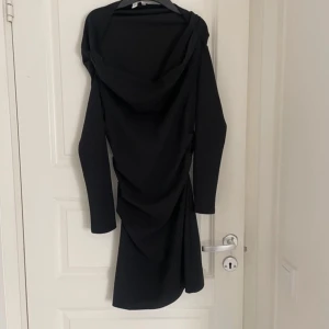 Svart asymmetrisk kortklänning - Snygg svart kortklänning med lång ärm och draperad design. Klänningen har en cool asymmetrisk form och en bred krage som ger en unik look. Perfekt för dig som vill sticka ut med en stilren och modern outfit.