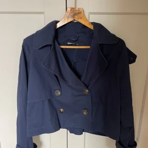 Mörkblå trenchcoat från Gina Tricot - Snygg mörkblå skepparkavaj från Gina Tricot i kortare modell. Jackan har dubbelknäppning med stora knappar, bred krage och långa ärmar. Perfekt för dig som vill ha en stilren och trendig look till vardags. Passar dig som gillar klassiska detaljer och enkel design.