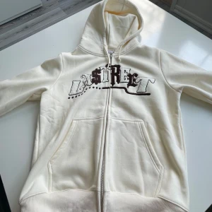 Hoodie från Lastreet - Säljer en krämvit hoodie från Lastreet med text i brunt och vitt på bröstet. Tröjan har dragkedja, huva med snörning och två stora fickor framtill. Perfekt för en chill streetstyle-look.